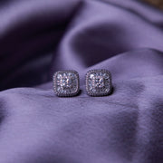 A pair of sterling silver stud earrings with a brilliant-cut white stone, set within a sterling silver bezel, displayed on a purple fabric surface.